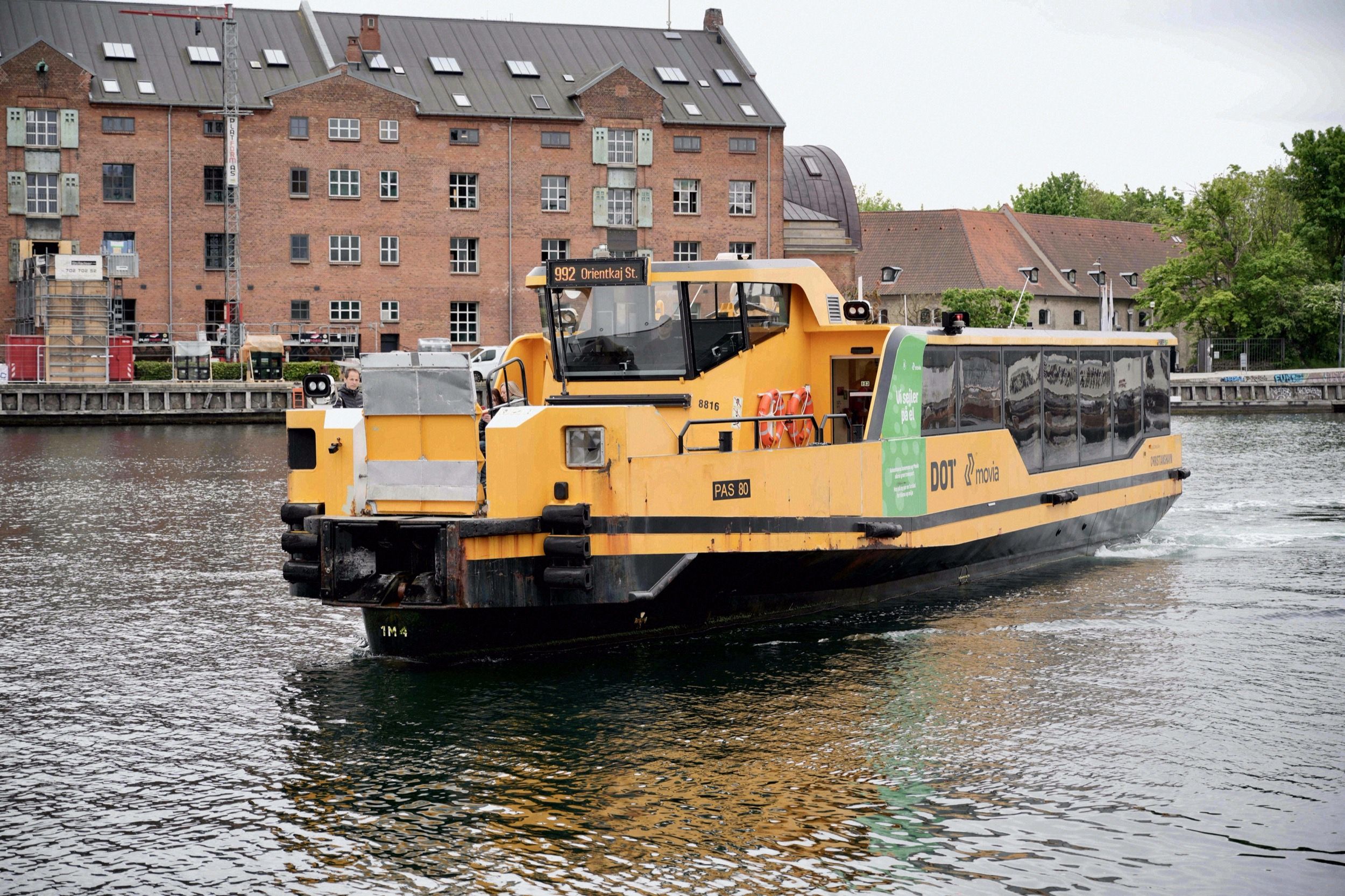 Der gelbe Kopenhagener Hafenbus (DOT Linie 992) faehrt auf dem Kanal in Christianshavn, flankiert von historischen Backsteinlagerhaeusern.