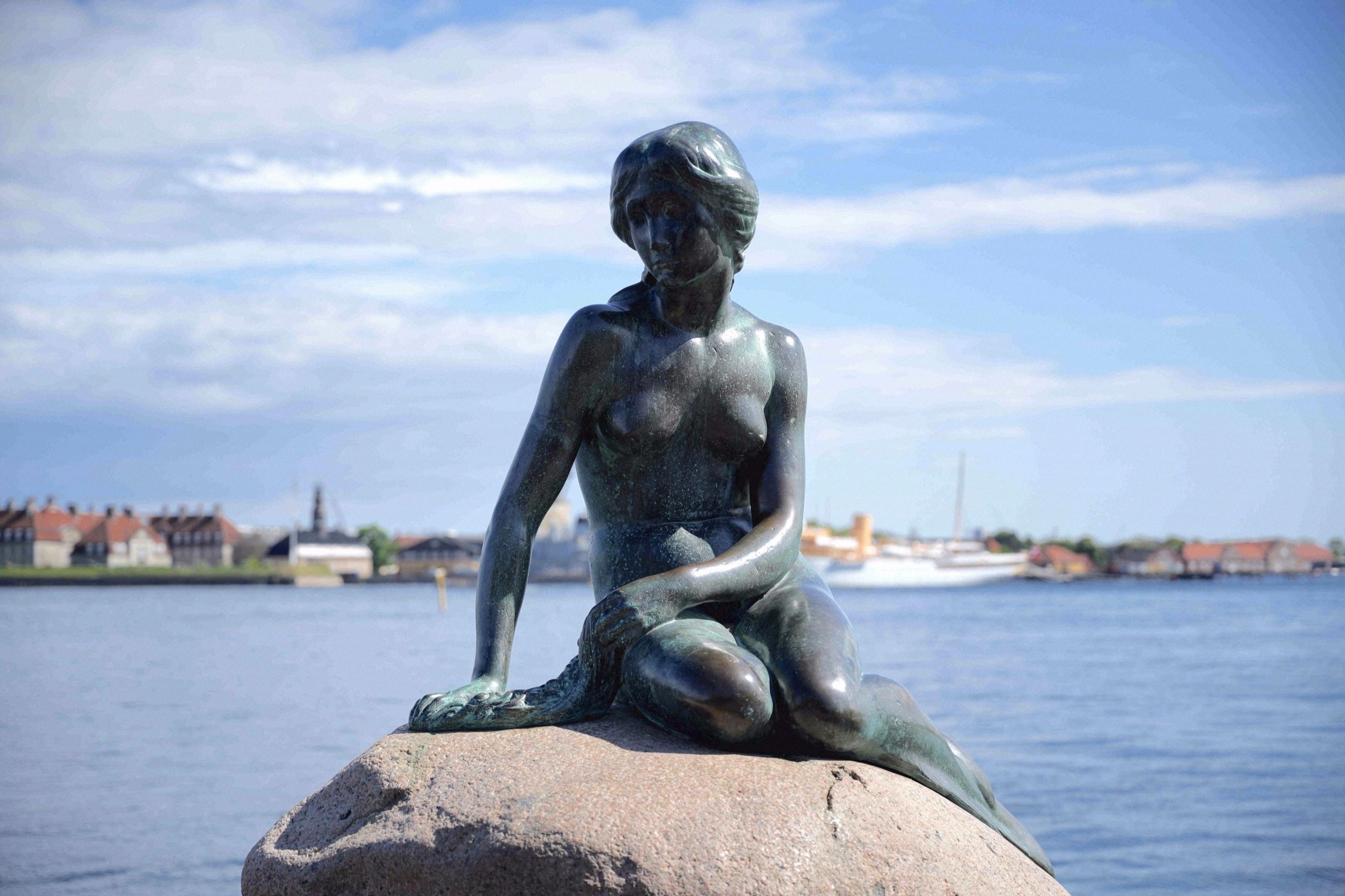 Die beruehmte Bronzestatue der Kleinen Meerjungfrau sitzt auf einem Felsen am Kopenhagener Hafen mit dem Stadtpanorama im Hintergrund.