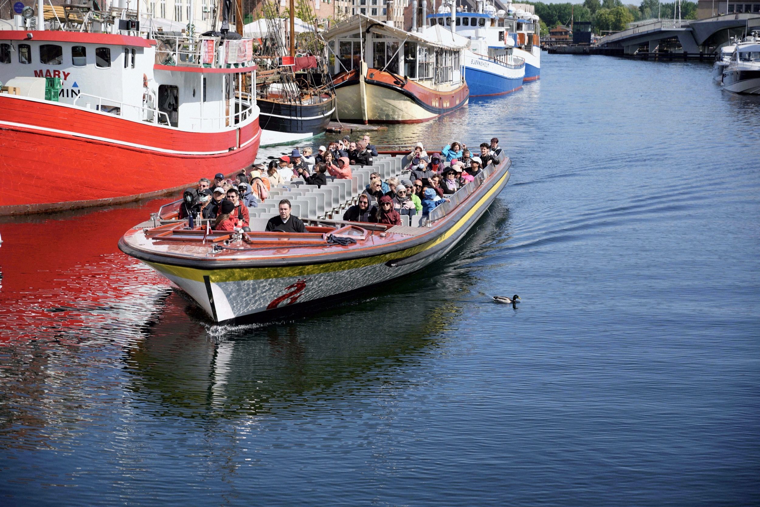 Ein vollbesetztes Kanaltour-Ausflugsboot faehrt am Nyhavn-Becken vorbei, im Hintergrund historische Hafen- und Museumsschiffe.