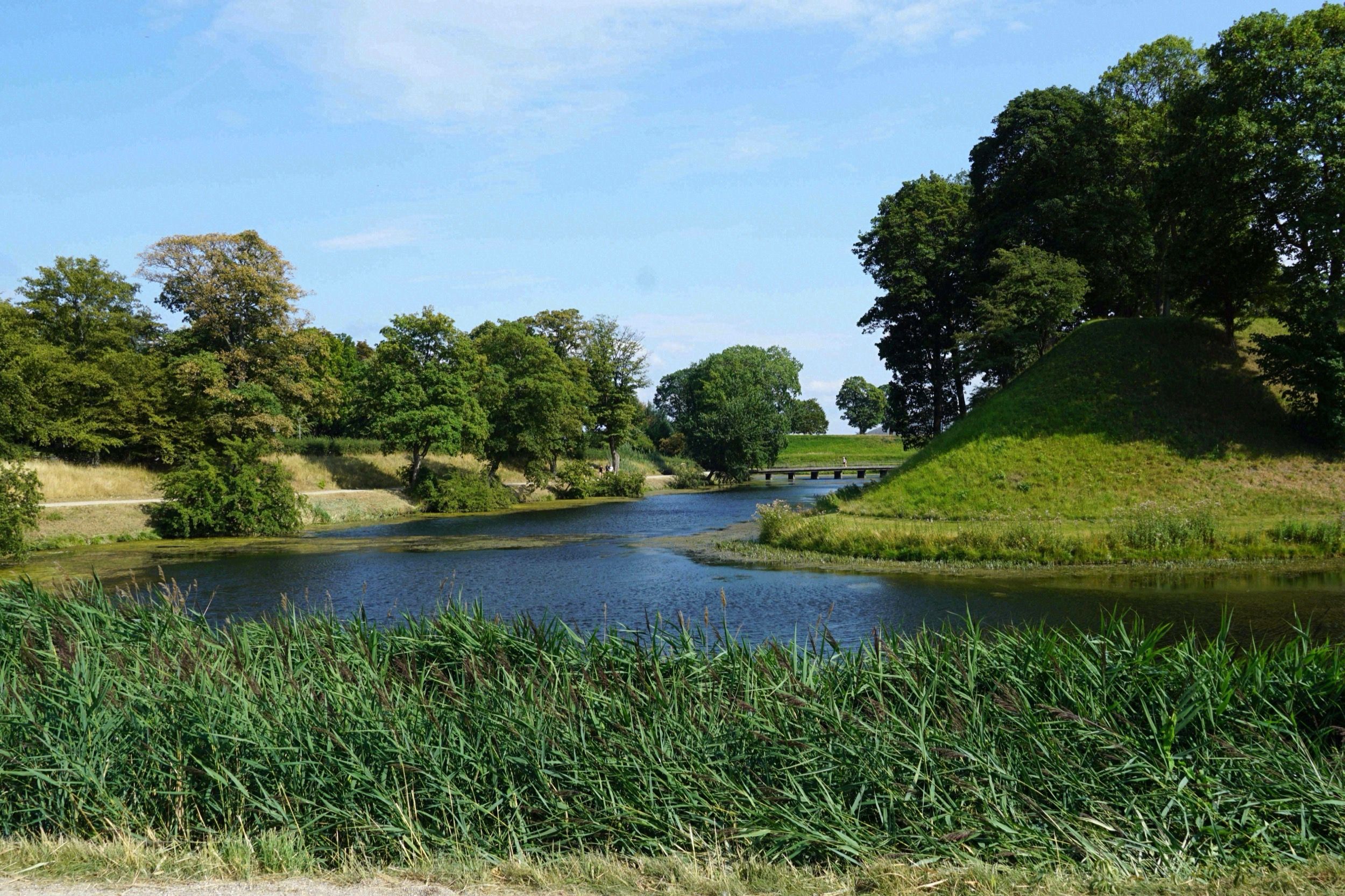 Wassergraben und begraster Erdwall der Sternfestung Kastellet mit einer kleinen Bruecke im Sommer.