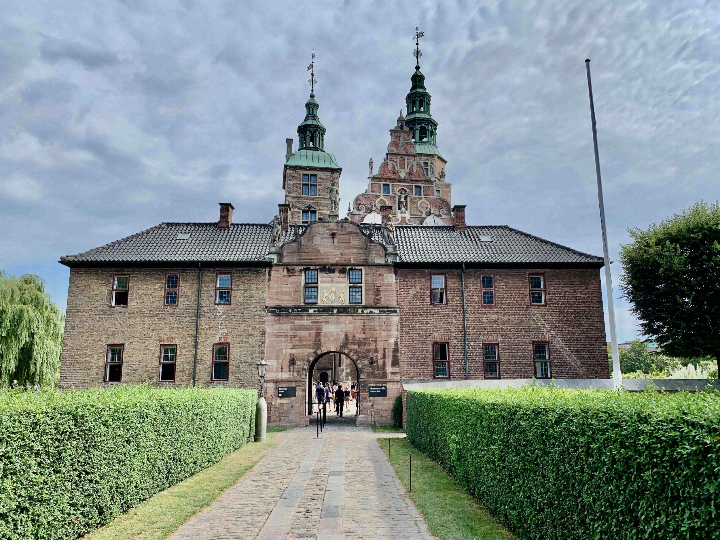 Frontansicht von Rosenborg Slot mit symmetrischem Toreingang, zwei gruenen Tuermschen und gepflegten Heckenrabatten im Vordergrund.