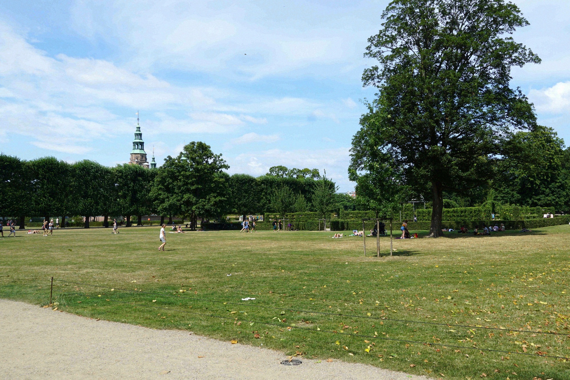Rosenborg Slot erhebt sich hinter alten Baeumen im Kongens Have, umgeben von Besuchern an einem sonnigen Sommertag.